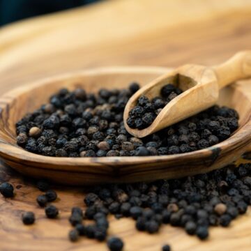 whole black peppers on a wooden tray with a wooden spoon.