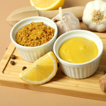 Small bowls of mustard and lemon slices and garlic bulbs on a wooden board.
