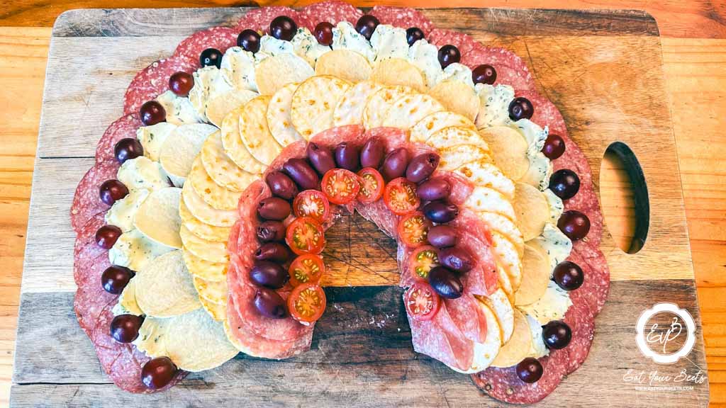 Decorating pepperoni, cream cheese, grapes, pringles, rice crackers, salami, cherry tomatoes, and olives on wooden board.