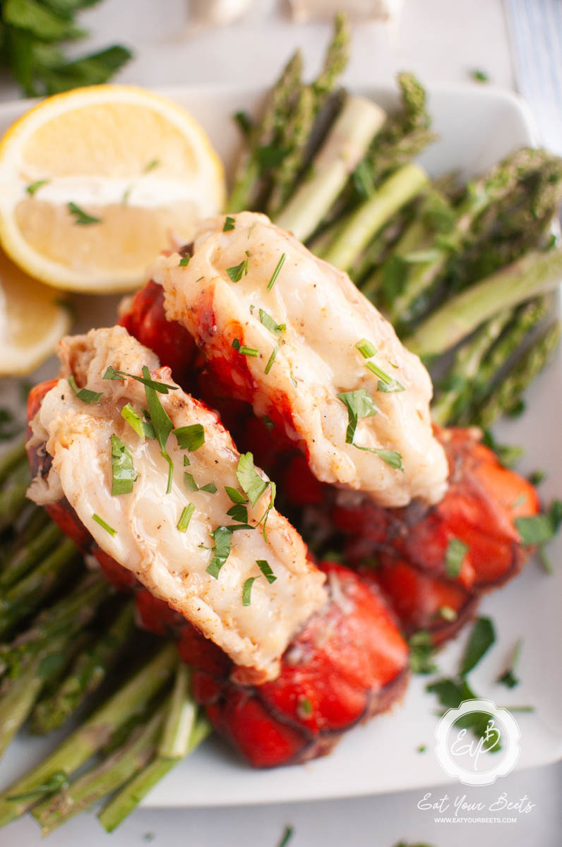 Sering garlic lobster tails with steamed asparagus and lemon wedges on a white plate.